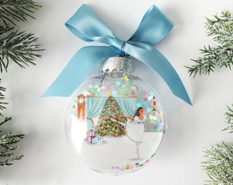 May include: A clear glass Christmas ornament with a blue ribbon bow. The ornament features a printed image of a ballerina standing in front of a decorated Christmas tree. The image is surrounded by iridescent glitter.