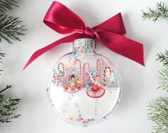May include: A clear glass Christmas ornament with a red bow and two ballerina figures inside. The ornament is filled with colorful confetti and candy canes.