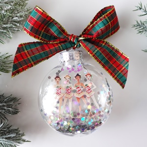 May include: A clear glass Christmas ornament with a red and green plaid ribbon bow. Inside the ornament are four figures of ballerinas wearing white tutus and red and white striped tops. The ornament is filled with iridescent glitter.