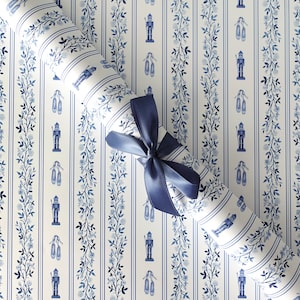 Puede incluir: Un rollo de papel de regalo a rayas azules y blancas con un lazo azul marino. El papel presenta un patrón de cascanueces, zapatillas de ballet y guirnaldas florales. El fondo es blanco con rayas azules verticales.