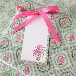 May include: A wrapped gift with floral patterned paper in shades of green and pink. A bright pink ribbon is tied in a bow, and a blank white gift tag with a pink floral design is attached.