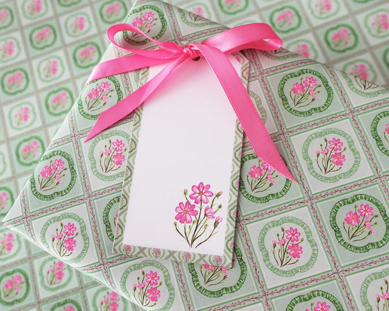 May include: A wrapped gift with floral patterned paper in shades of green and pink. A bright pink ribbon is tied in a bow, and a blank white gift tag with a pink floral design is attached.