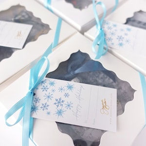 K&ouml;nnte beinhalten: Wei&szlig;e Geschenkboxen mit blauen Schleifen und Schneeflocken-Geschenk-Etiketten. Die Etiketten sagen "Frohe Feiertage" und haben einen Platz zum Schreiben von "An :" und "Von :"