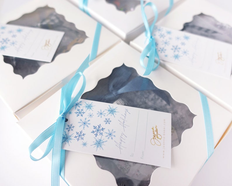 K&ouml;nnte beinhalten: Wei&szlig;e Geschenkboxen mit blauen Schleifen und Schneeflocken-Geschenk-Etiketten. Die Etiketten sagen "Frohe Feiertage" und haben einen Platz zum Schreiben von "An :" und "Von :"
