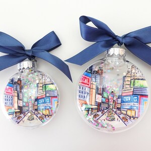 K&ouml;nnte beinhalten: Zwei transparente Weihnachtskugeln aus Glas mit einer Stadtlandschaft im Inneren. Die Kugeln sind mit Glitzer verziert und haben blaue Schleifen.