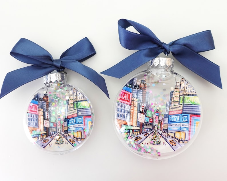 K&ouml;nnte beinhalten: Zwei transparente Weihnachtskugeln aus Glas mit einer Stadtlandschaft im Inneren. Die Kugeln sind mit Glitzer verziert und haben blaue Schleifen.