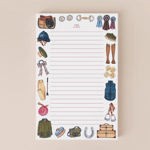 May include: A lined notepad with equestrian-themed illustrations. The notepad features images of riding equipment, including a saddle, helmet, boots, and a horseshoe. The initials "JBL" are printed at the top. The notepad is surrounded by a light beige background.