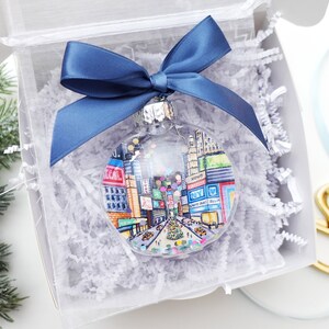 K&ouml;nnte beinhalten: Ein transparenter Weihnachtsbaumschmuck aus Glas mit einer handgemalten Illustration des Times Square in New York City. Der Schmuck ist mit bunten Konfetti und einem blauen Band verziert.