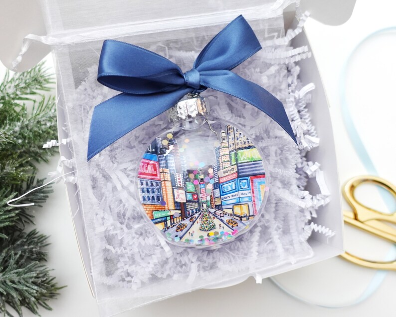 K&ouml;nnte beinhalten: Ein transparenter Weihnachtsbaumschmuck aus Glas mit einer handgemalten Illustration des Times Square in New York City. Der Schmuck ist mit bunten Konfetti und einem blauen Band verziert.