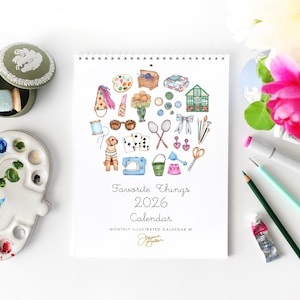 Może przedstawiać: Spiralny kalendarz zatytułowany "Favorite Things 2026 Calendar" z akwarelowymi ilustracjami różnych przedmiotów. Paleta malarska, pędzle i materiały artystyczne są ułożone wokół kalendarza. Obecne jest również ozdobne pudełko i kwiaty.