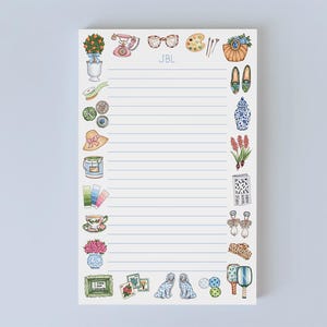 May include: A lined notepad with decorative illustrations of various objects, including a telephone, glasses, a teacup, and a hat. The notepad has the initials "JBL" at the top. The illustrations are in a watercolor style with pastel colors.