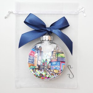 K&ouml;nnte beinhalten: Ein transparenter Weihnachtsbaumschmuck aus Glas mit einer handgemalten Illustration des Times Square in New York City. Der Schmuck ist mit buntem Glitzer gef&uuml;llt und wird mit einem silbernen Aufh&auml;nger geliefert.