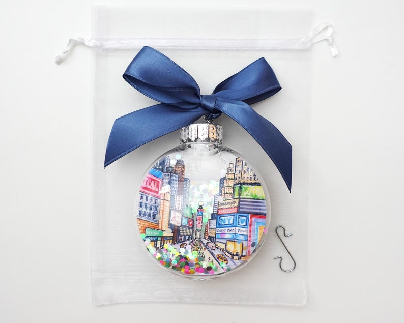 K&ouml;nnte beinhalten: Ein transparenter Weihnachtsbaumschmuck aus Glas mit einer handgemalten Illustration des Times Square in New York City. Der Schmuck ist mit buntem Glitzer gef&uuml;llt und wird mit einem silbernen Aufh&auml;nger geliefert.