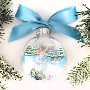 May include: A clear Christmas ornament with a ballerina design. The ornament features a ballerina in a white tutu, dancing in a winter scene with snow-covered trees. A light blue ribbon is tied at the top. Green pine branches surround the ornament.