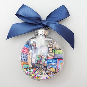 K&ouml;nnte beinhalten: Ein transparenter Weihnachtsbaumschmuck aus Glas mit einer handgemalten Szene des Times Square in New York City. Der Schmuck ist mit buntem Glitzer gef&uuml;llt und hat ein blaues Band, das um die Oberseite gebunden ist.
