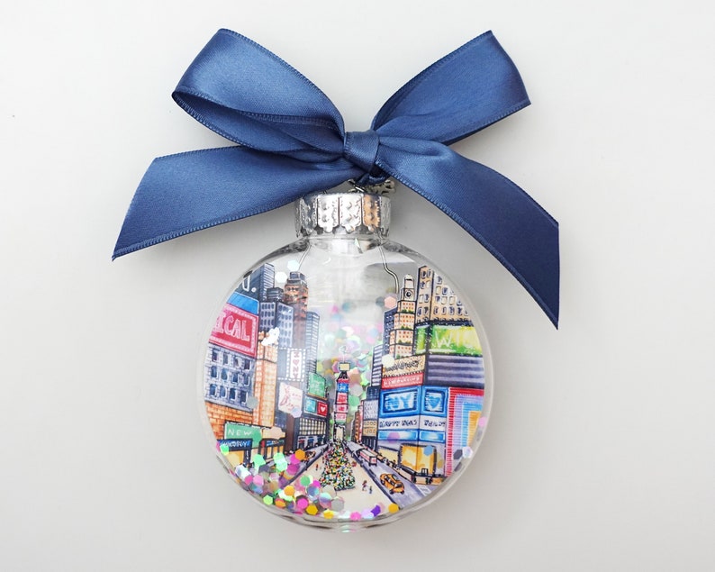 K&ouml;nnte beinhalten: Ein transparenter Weihnachtsbaumschmuck aus Glas mit einer handgemalten Szene des Times Square in New York City. Der Schmuck ist mit buntem Glitzer gef&uuml;llt und hat ein blaues Band, das um die Oberseite gebunden ist.