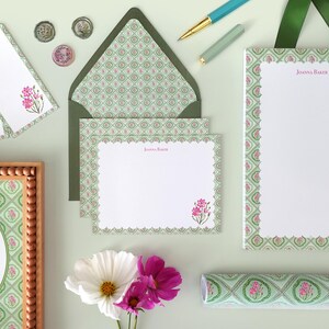 May include: Stationery set featuring a floral design with pink flowers and a green and white pattern. Includes notepaper, envelopes, tags, and a framed print. A pen and ribbon are also visible.