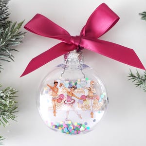 May include: A clear glass Christmas ornament with a pink ribbon. Inside the ornament are three ballerinas with colorful confetti.
