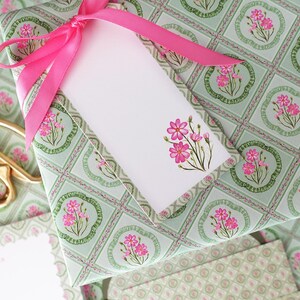May include: A wrapped gift with a pink ribbon and a white gift tag. The wrapping paper has a light green background with a floral pattern. Gold scissors are visible in the background.