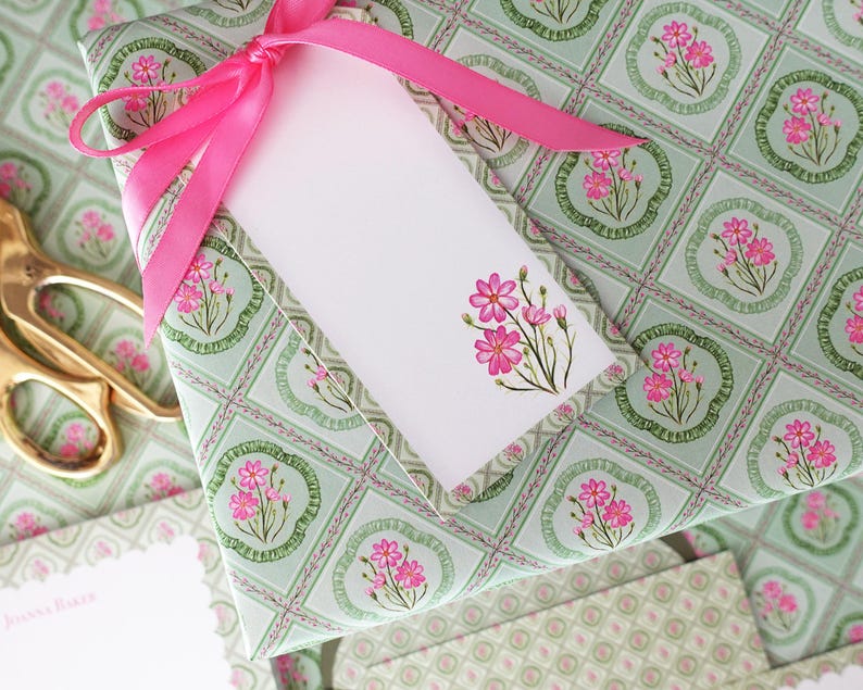 May include: A wrapped gift with a pink ribbon and a white gift tag. The wrapping paper has a light green background with a floral pattern. Gold scissors are visible in the background.