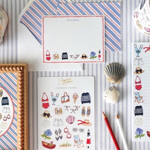K&ouml;nnte beinhalten: Eine Auswahl an nautisch thematisierter Schreibwaren und Aufklebern. Das Bild zeigt einen gerahmten Druck, Notizbl&ouml;cke, Aufkleber und eine Tasse, alle mit Illustrationen von Strandartikeln wie Badeanz&uuml;gen, H&uuml;ten und Segelbooten. Die Farbpalette ist haupts&auml;chlich rot, wei&szlig; und blau.