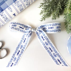 May include: A white ribbon with a blue pattern featuring nutcrackers, ballet slippers, and other decorative elements. The ribbon is tied into a bow, with rolls of matching wrapping paper and scissors in the background.