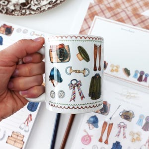 Puede incluir: Taza de cerámica blanca con ilustraciones de temática ecuestre, que incluyen una silla de montar, botas de montar y un casco de montar. La taza tiene un borde decorativo y se sostiene cerca de artículos de papelería con diseños similares. El tema es ecuestre.