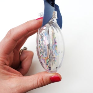 K&ouml;nnte beinhalten: Ein transparenter Glas-Weihnachtsbaumschmuck gef&uuml;llt mit bunten Konfetti und einem gedruckten Bild einer Stadtlandschaft. Der Schmuck hat eine silberne Spitze und h&auml;ngt an einem blauen Band.