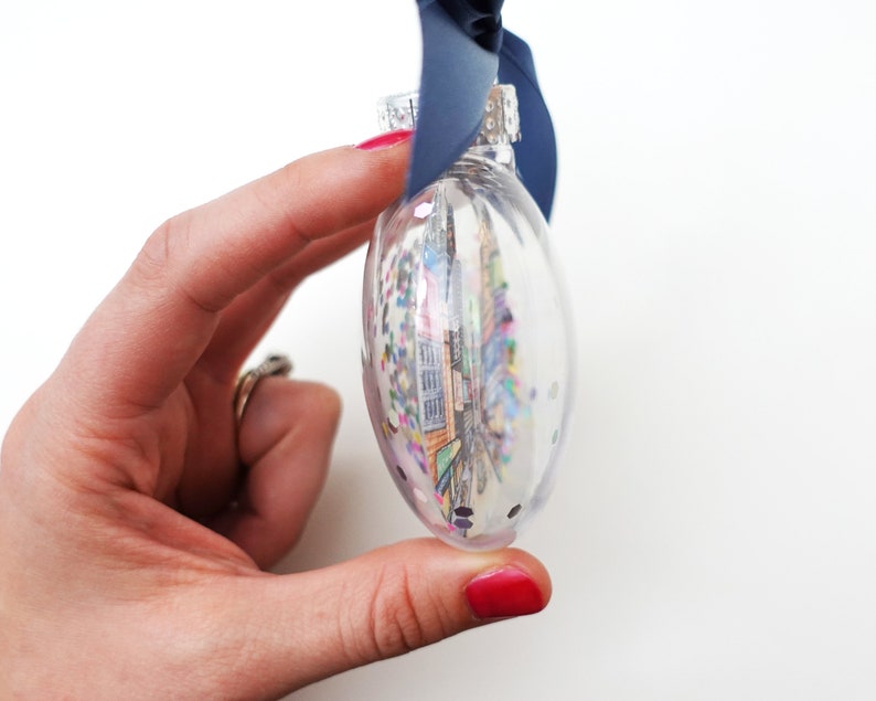 K&ouml;nnte beinhalten: Ein transparenter Glas-Weihnachtsbaumschmuck gef&uuml;llt mit bunten Konfetti und einem gedruckten Bild einer Stadtlandschaft. Der Schmuck hat eine silberne Spitze und h&auml;ngt an einem blauen Band.