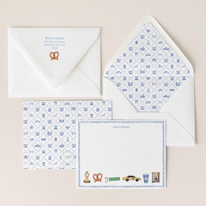 May include: A stationery set featuring white envelopes and note cards. One envelope shows the text Joanna Baker, 225 Bennett Avenue, New York, New York 10040 with a pretzel icon. The cards and envelope liners feature a blue and white pattern of New York City icons. A note card displays a blue border and illustrations of a clock, pretzel, street sign, taxi, coffee cup, and building.