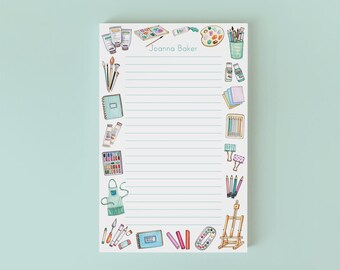 May include: A lined notepad with a light blue background. The notepad is decorated with watercolor illustrations of art supplies, including paint tubes, brushes, a palette, and an easel. The name "Joanna Baker" is printed at the top.