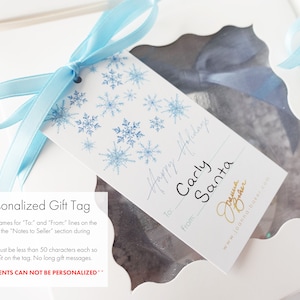K&ouml;nnte beinhalten: Ein wei&szlig;es Geschenkanh&auml;nger mit einem blauen Schneeflockenmuster. Der Anh&auml;nger hat den Text "Happy Holidays" in Kursivschrift. Der Anh&auml;nger hat auch den Text "To: Carly Santa" in Kursivschrift. Der Anh&auml;nger hat den Text "From:" in Kursivschrift. Der Anh&auml;nger hat den Text "Joanna Baker" in Kursivschrift. Der Anh&auml;nger hat den Text "www.joanna-baker.com" in Kursivschrift.