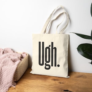 May include: Cream-colored tote bag with the word "Ugh." printed in bold, black letters. The bag has a simple design with long handles. The tote bag is made of a light-colored fabric and is sitting on a wooden surface.