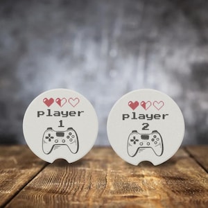 May include: Two white ceramic coasters with a red heart graphic and the text "Player 1" and "Player 2" with a video game controller graphic.