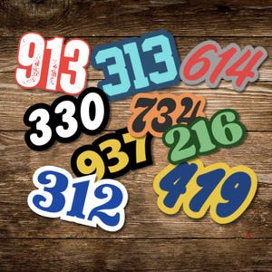May include: A collection of colorful stickers featuring various area code numbers, including 913, 313, 614, 330, 732, 216, 937, 312, and 419. The stickers are in different colors and fonts, set against a wooden background.