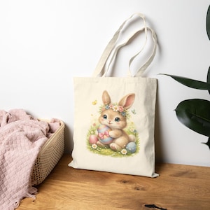 May include: A beige tote bag featuring a watercolor illustration of a cute bunny wearing a flower crown and holding a decorated Easter egg. The bunny is surrounded by colorful eggs and flowers, with butterflies fluttering around.