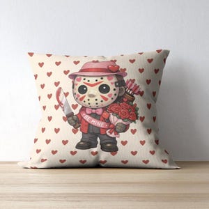 Può includere: Un cuscino decorativo con un personaggio dei cartoni animati con una maschera da hockey, che tiene un coltello e un mazzo di rose. Il personaggio indossa un cappello e una giacca rosa, con le parole "BE MINE" su uno striscione. Il cuscino ha uno sfondo crema con cuori rossi.