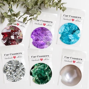 May include: A collection of car coasters with various gemstone designs, including red, purple, blue, silver, and green. Each coaster is packaged in a clear plastic sleeve. The text "Car Coasters Handmade with love" is printed on each package.