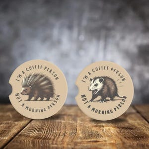 May include: Two round brown coasters with a white border. The coasters feature a black and white illustration of an animal. The text on the coasters reads "I'm a coffee person, not a morning person".