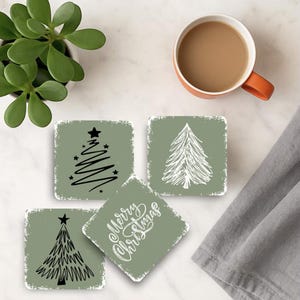 May include: Four square coasters with a distressed sage green background and white and black Christmas tree designs. One coaster has the words "Merry Christmas" in white script. A cup of coffee and a plant are also in the image.