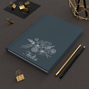 May include: A dark blue notebook with a white floral design and the word "Notes" printed on the cover. Two pencils are lying on a dark surface next to the notebook.