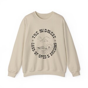 May include: Cream-colored sweatshirt with a crew neck. The front features a circular graphic with the text "The Midwest Land of Hopes & Dreams" in a black, stylized font. The graphic includes illustrations of a farm, fields, clouds, and a sun.