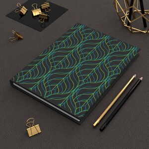 May include: A black notebook with a green and blue geometric leaf pattern. The notebook is lying on a dark gray surface with two pencils and gold paper clips.