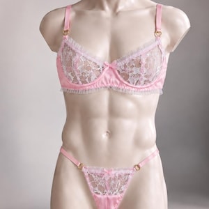 Può includere: Un completo di lingerie in pizzo rosa e bianco composto da un reggiseno con ferretto e un perizoma abbinato. Il reggiseno presenta spalline in raso rosa, anelli dorati e coppe in pizzo bianco con bordo a volant rosa e un fiocchetto centrale.