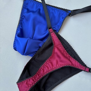 Puede incluir: Dos conjuntos de lencería. Uno es azul vibrante, el otro burdeos oscuro. Ambos tienen ribetes y tirantes negros. Los bordes están acabados con un delicado encaje festoneado. La tela parece ser un satén liso.