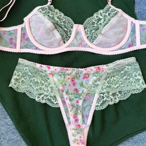 May include: A delicate lingerie set featuring a floral print. The bra and matching underwear are trimmed with light green lace and pink accents. The bra has adjustable straps. The set is displayed on a dark green surface.