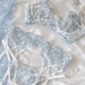 K&ouml;nnte beinhalten: Zartes Dessous-Set in Hellblau und Wei&szlig; mit floralen Spitzendetails. Das Set umfasst einen BH, einen Strumpfhalter und einen Slip, alles auf einer wei&szlig;en Satin-Oberfl&auml;che angeordnet. Verstreute Perlen sorgen f&uuml;r einen Hauch von Eleganz.