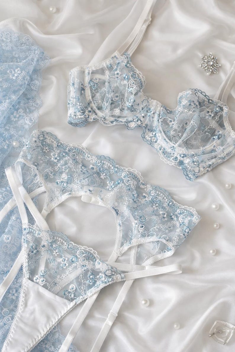 K&ouml;nnte beinhalten: Zartes Dessous-Set in Hellblau und Wei&szlig; mit floralen Spitzendetails. Das Set umfasst einen BH, einen Strumpfhalter und einen Slip, alles auf einer wei&szlig;en Satin-Oberfl&auml;che angeordnet. Verstreute Perlen sorgen f&uuml;r einen Hauch von Eleganz.