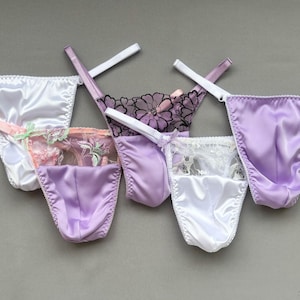 May include: Assortment of satin thong underwear in white, lavender, and purple. Some feature lace or floral accents. The lingerie is displayed on a gray background, showcasing the delicate fabrics and designs.