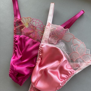 Pode incluir: Duas peças de lingerie de cetim rosa. Uma é fúcsia vibrante, a outra rosa pálido. Ambas apresentam delicados detalhes de renda floral e alças ajustáveis. As bordas são finalizadas com uma guarnição decorativa.